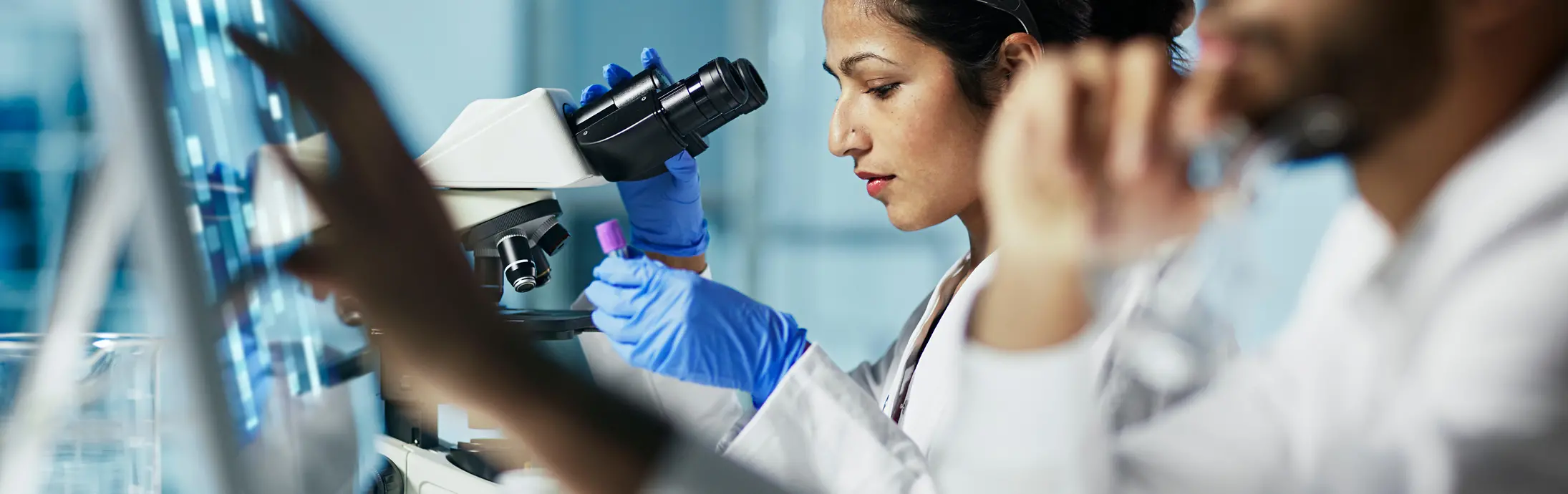 laboratorija s ženom koja sedi ispred mikroskopa, posmatra uzorak, i ispred nje muškarac s bradom gleda u ekran