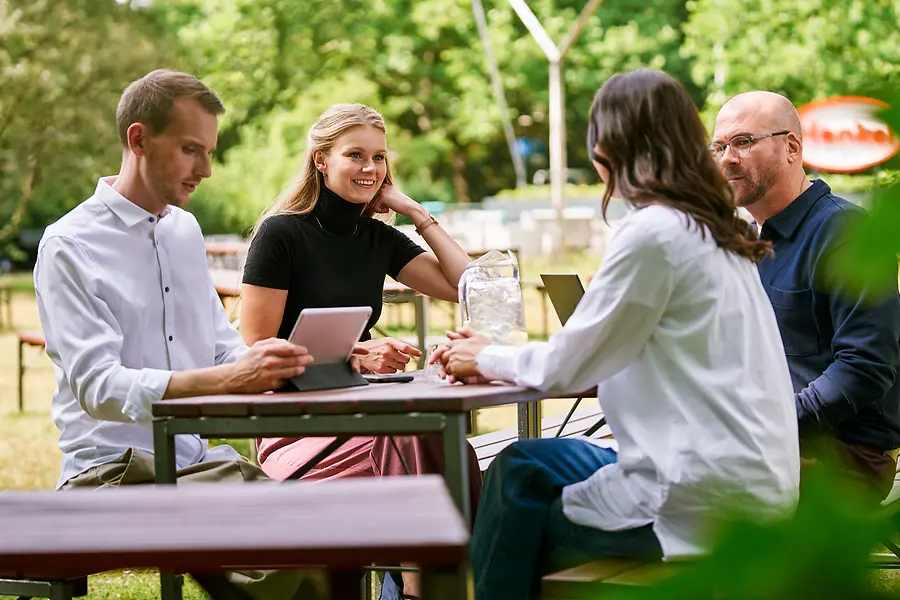 Grupa kolega sedi zajedno na otvorenom i razgovara o poslu uz tablete i laptopove, što predstavlja saradnju i otvorenu, timski orijentisanu kulturu kompanije Henkel.