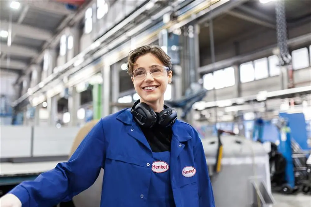 Nasmešeni zaposleni u Henkelu u plavom radnom mantilu i sa zaštitnim slušalicama stoji u industrijskoj proizvodnoj hali i predstavlja stručnost i inovacije u proizvodnji.
