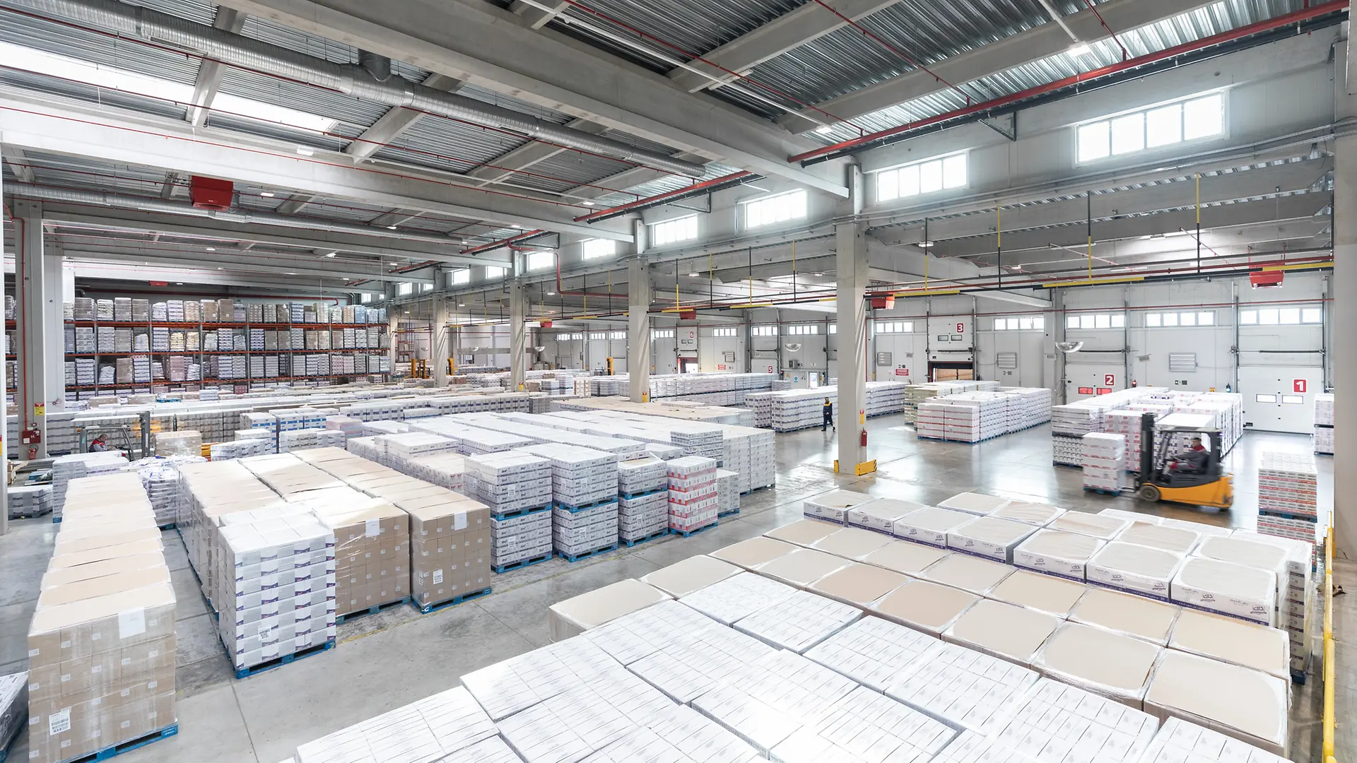 Na fotografiji je unutrašnjost velikog skladišta sa visokim policama i paletama uredno složenim po redovima. U prostoru se nalazi viljuškar koji pomera robu, a objekat je osvetljen prirodnim svetlom kroz prozore na krovu.