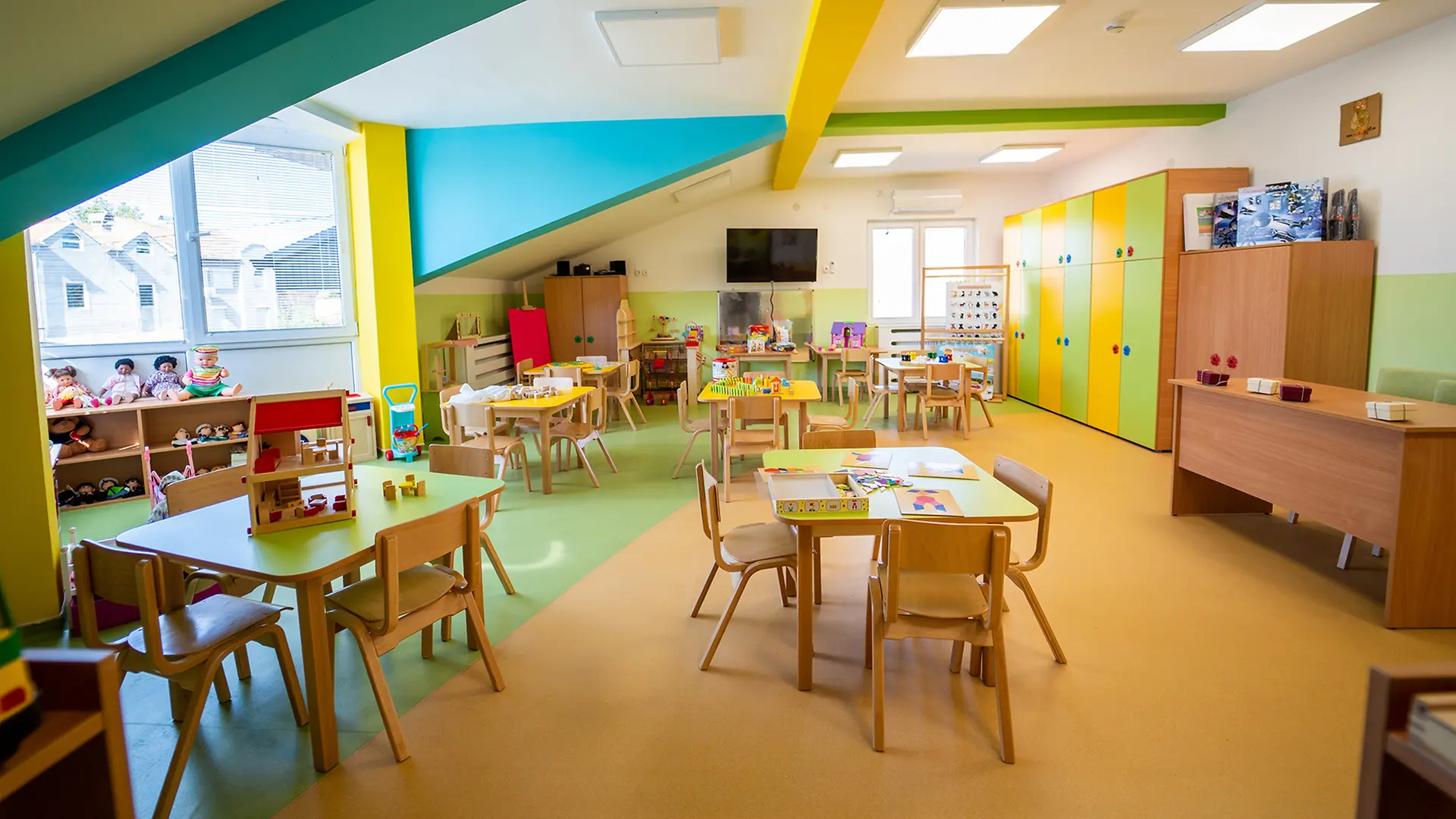Na fotografiji je svetla i šarena učionica u vrtiću „Čarolija“ u Svilajncu, opremljena drvenim stolovima i stolicama, igračkama, edukativnim materijalima i ormarima.