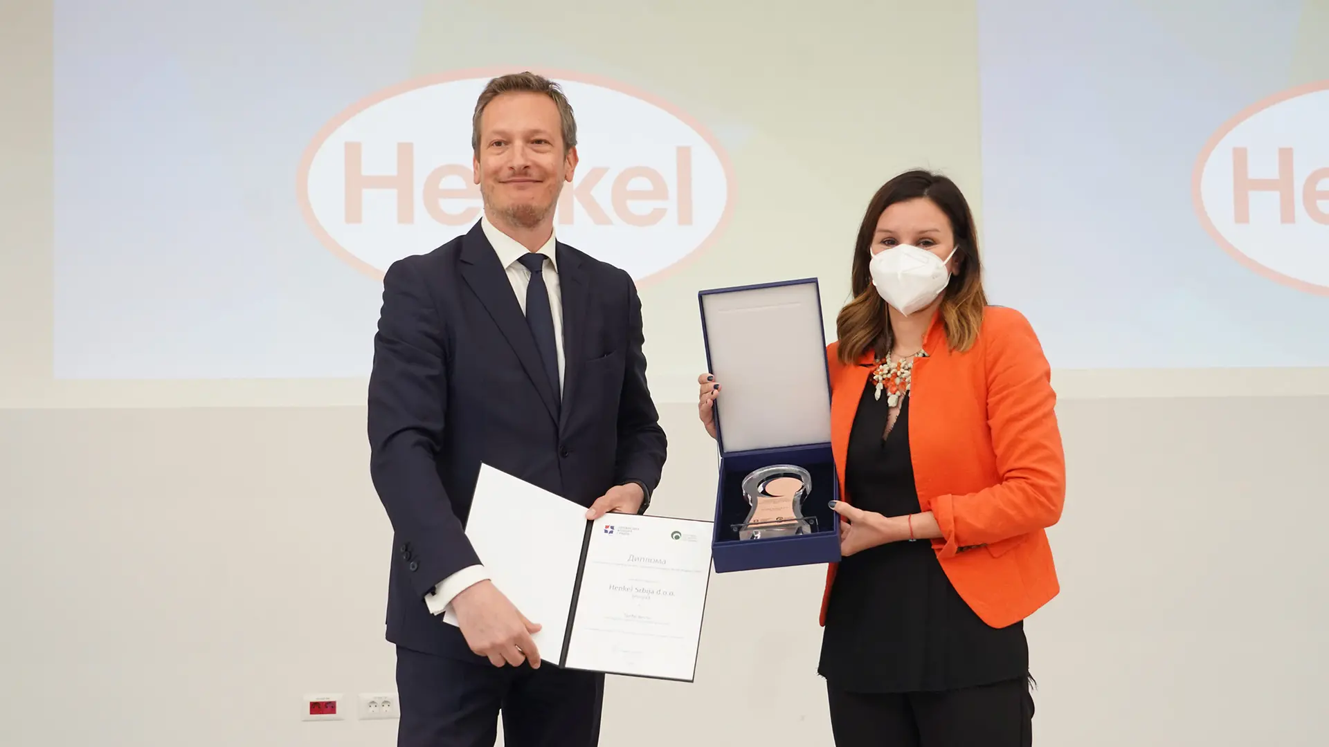 Na fotografiji je svečana dodela nagrade – prikazan je trenutak uručenja priznanja i sertifikata, uz Henkel logotip na ekranu u pozadini.