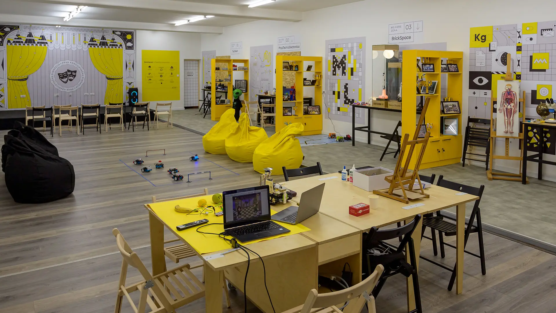 Na fotografiji je moderna učionica u okviru Mejkers lab projekta, opremljena stolovima, laptopom, edukativnim materijalima i žutim policama sa eksponatima. U prostoru se nalaze žuti i crni bean bag jastuci, kao i posteri sa naučnim temama na zidovima.