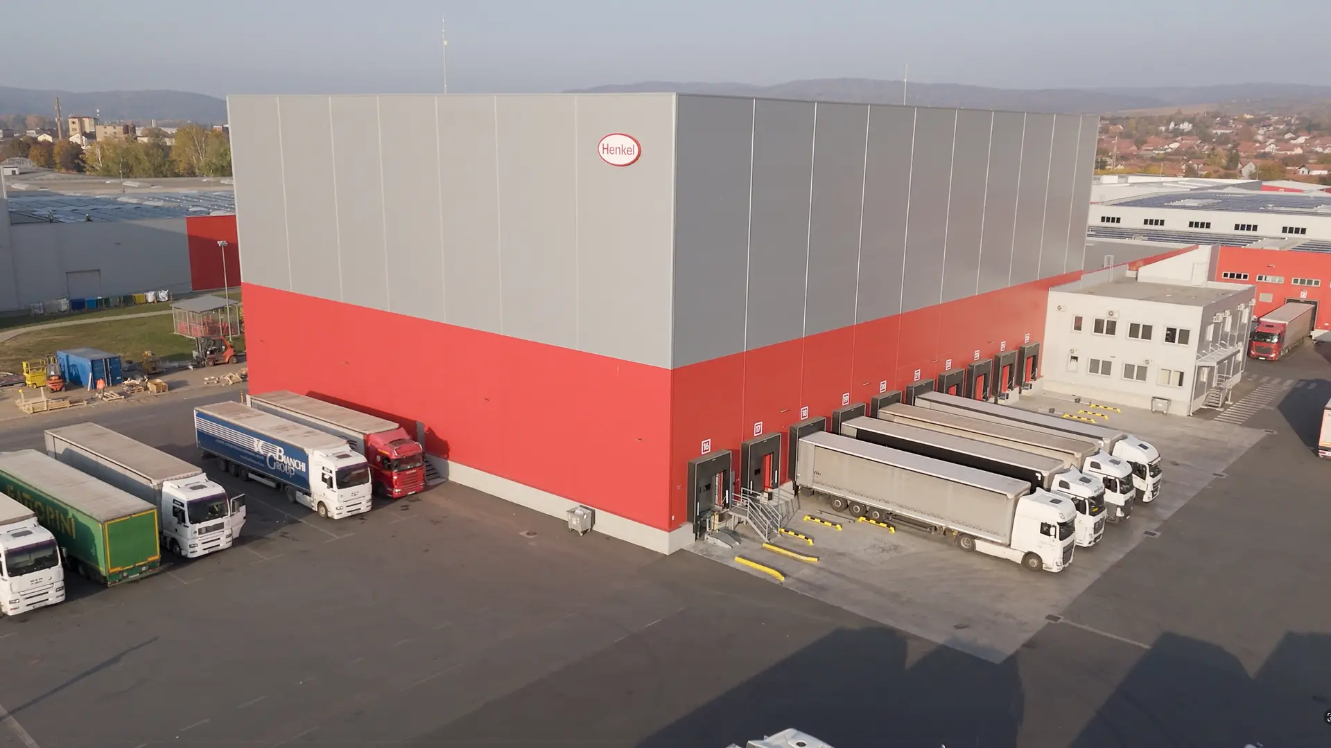 Na fotografiji je spoljašnji prikaz velikog Henkel skladišta u crveno-sivoj boji, sa više utovarnih rampi i parkiranim kamionima za transport robe.