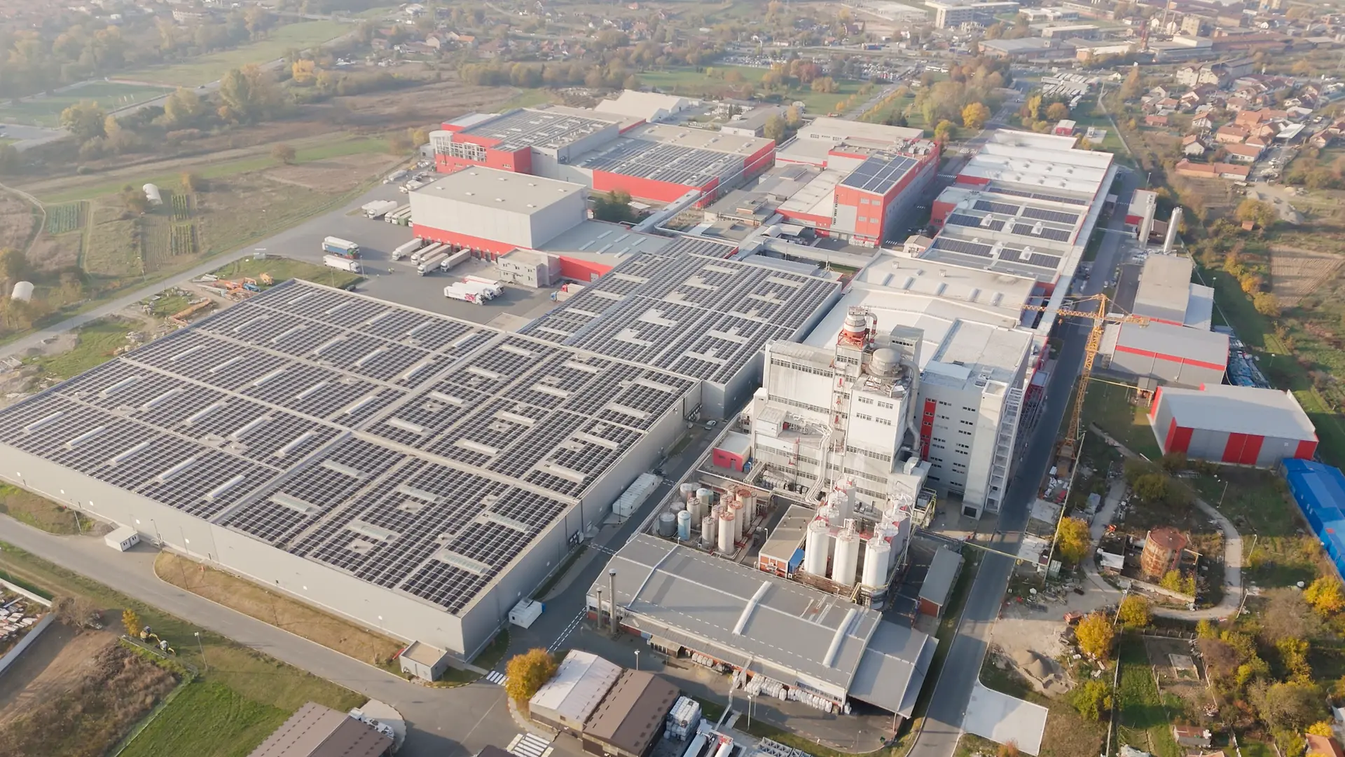 Na fotografiji je prikaz proizvodne lokacije Henkela u Kruševcu, sa brojnim objektima u crveno-sivoj boji. Na krovovima se nalaze solarni paneli, što ukazuje na završetak radova na solarnoj elektrani.