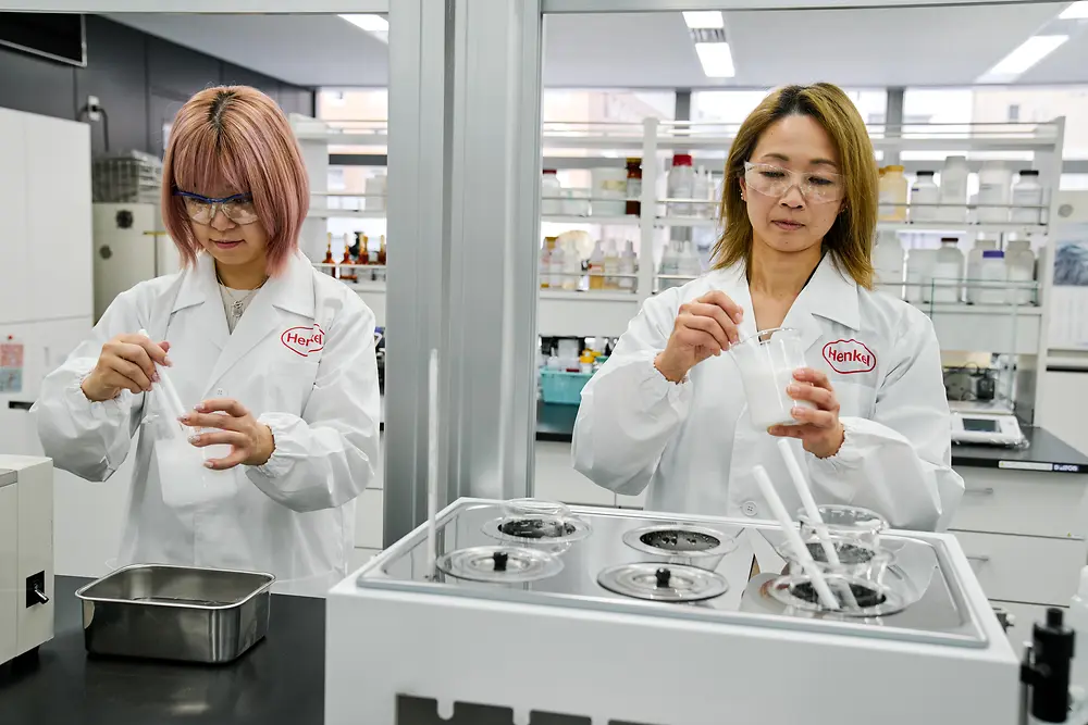 Dve osobe u belim Henkel laboratorijskim mantilima rade u modernoj laboratoriji. Rukuju naučnim instrumentima i posudama na radnoj površini, a u pozadini se vide različita laboratorijska oprema i police sa potrošnim materijalom.