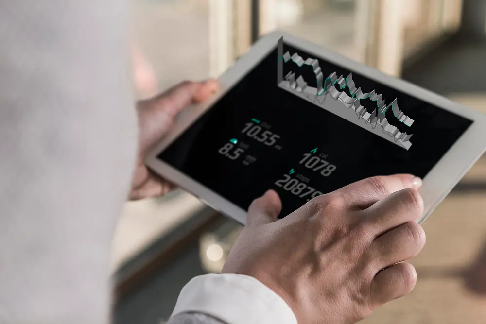Osoba sa tabletom pokazuje digitalne tabele i numeričke podatke, posmatrano preko nečijeg ramena u svetlom zatvorenom prostoru. 